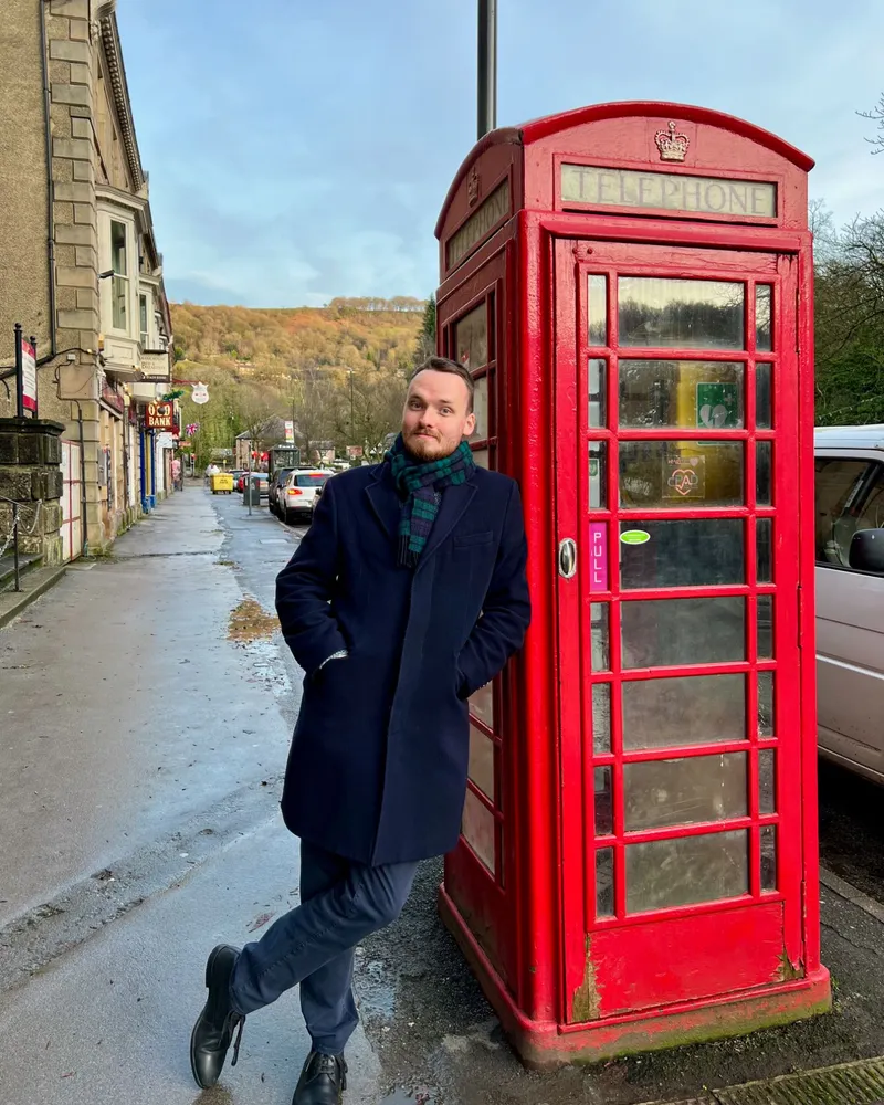 Anthony in a navy coat and tartan scarf, leaning on a red telephone box on a high street in Matlock, England